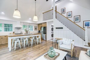 Eine geräumige Küche mit einer zentralen Kochinsel, Barhocker, einem Essbereich mit weißem Sofa und einer Treppe mit gerahmten Bildern.