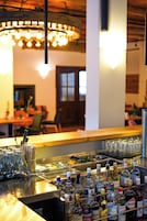Zona de bar con diversas botellas de licor y vasos, una lámpara colgante y un área de comedor al fondo.