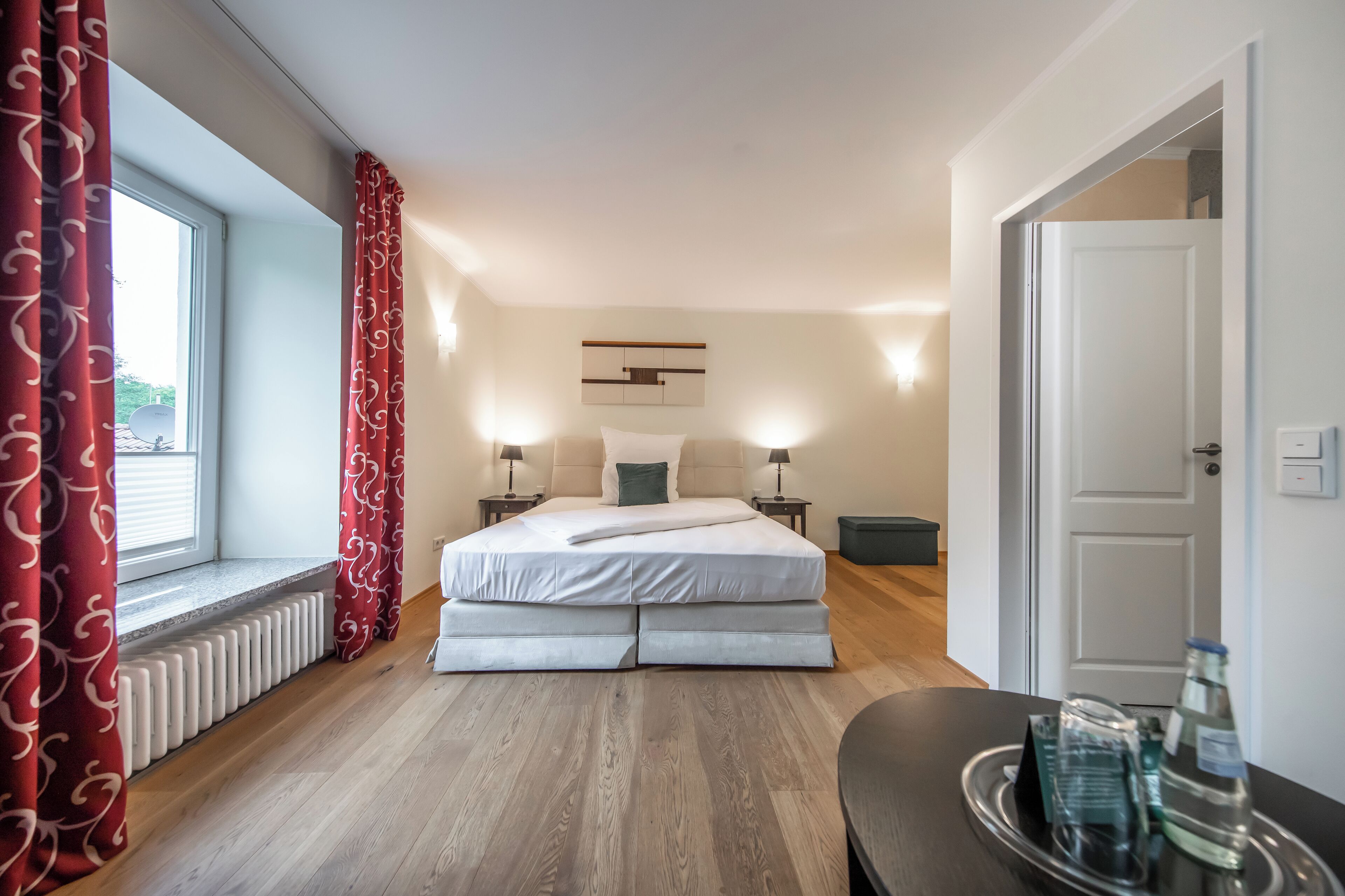 Une chambre à coucher avec un lit, des tables de chevet, un radiateur, une fenêtre avec des rideaux et une porte.