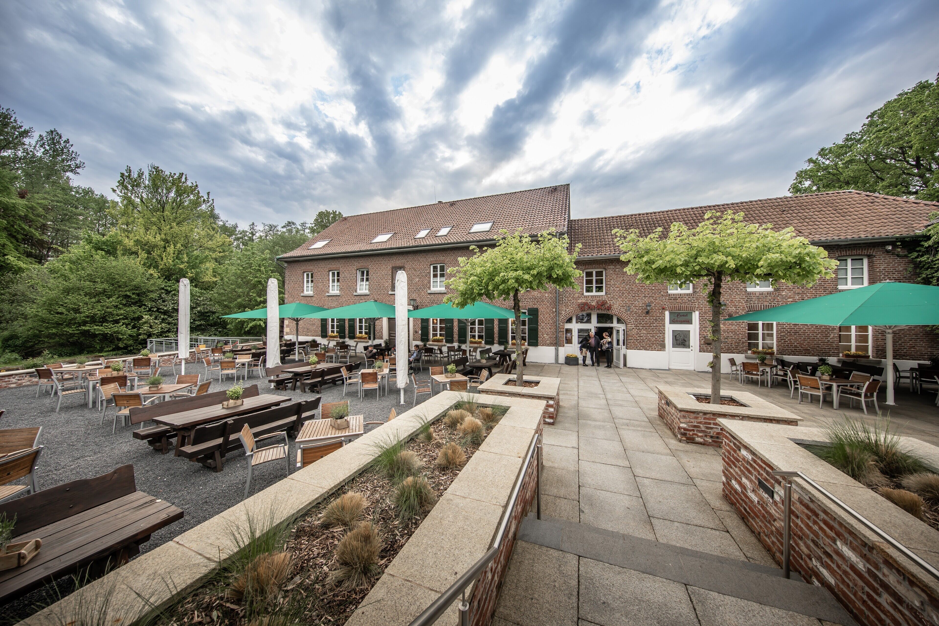 Een terras buiten met bakstenen gebouwen, groene parasols en houten tafels en stoelen.