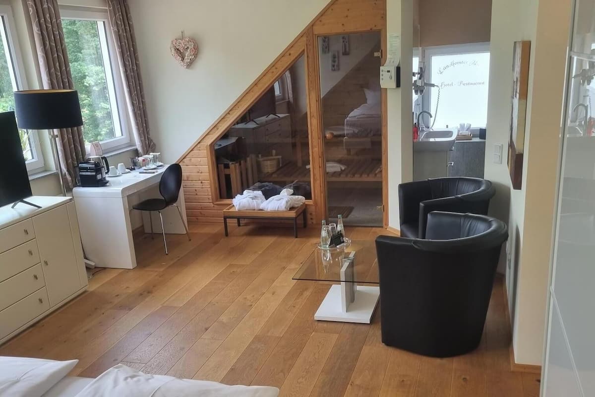 Una habitación de hotel moderna con piso de madera, una cama, un escritorio con silla, una mesita de noche con lámpara y un espejo grande.