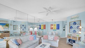 A beach-themed living room with wicker furniture, a ceiling fan, and wall decorations including framed pictures and a shelf with decorative plates. - The Palms 714 by Liquid Life (Orange Beach)