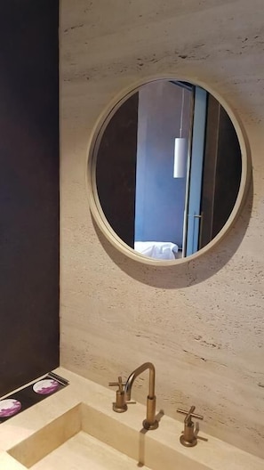 Kylpyhuoneessa on pyöreä peili, pesuallas, jossa on kaksi hanaa, ja pieni ikkuna.