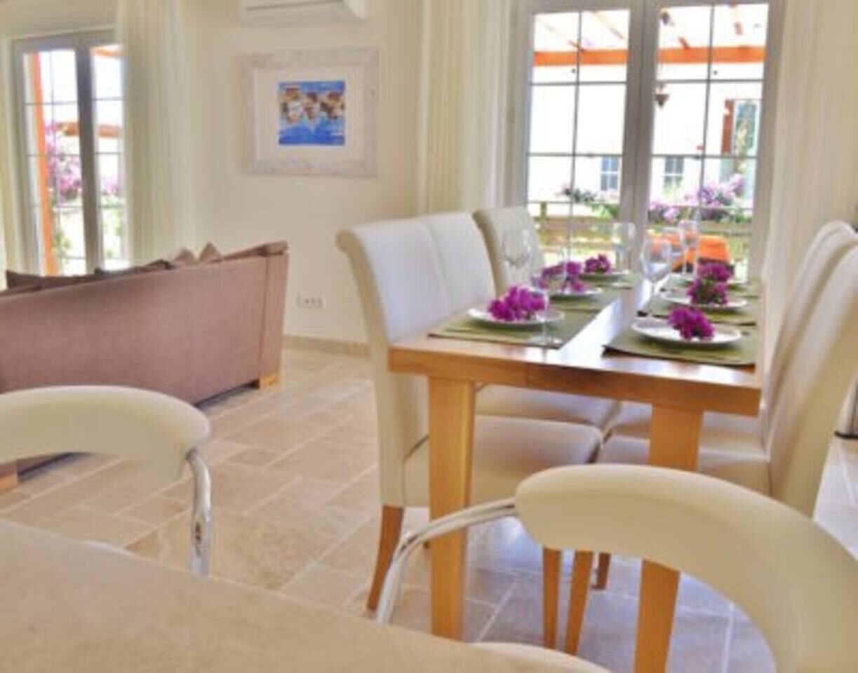 A dining area with a table set for four, chairs, a wooden table, and a view of the outdoors through large windows.