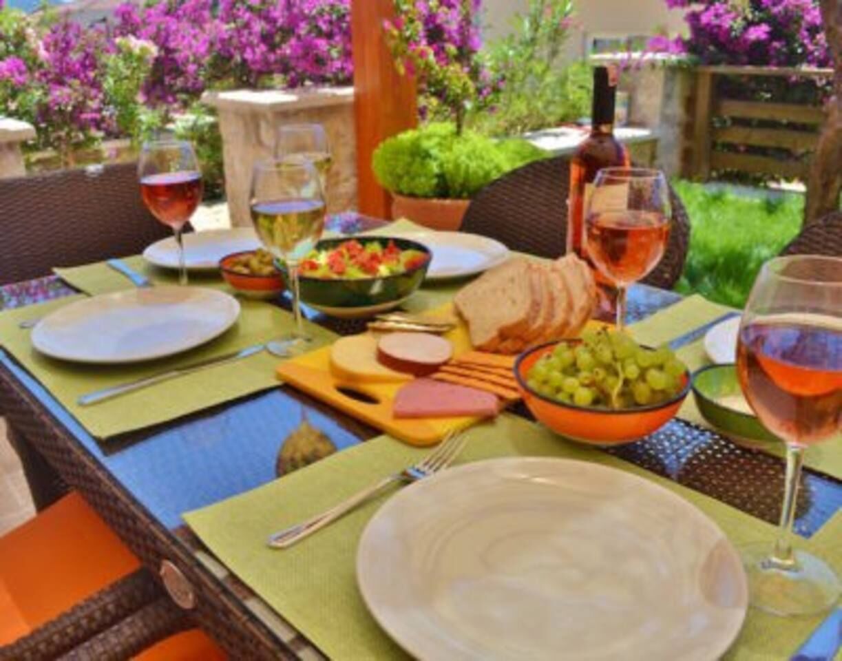 Une table dressée pour un dîner en plein air, avec de l’argenterie, des assiettes, un saladier de raisins et une bouteille de vin.