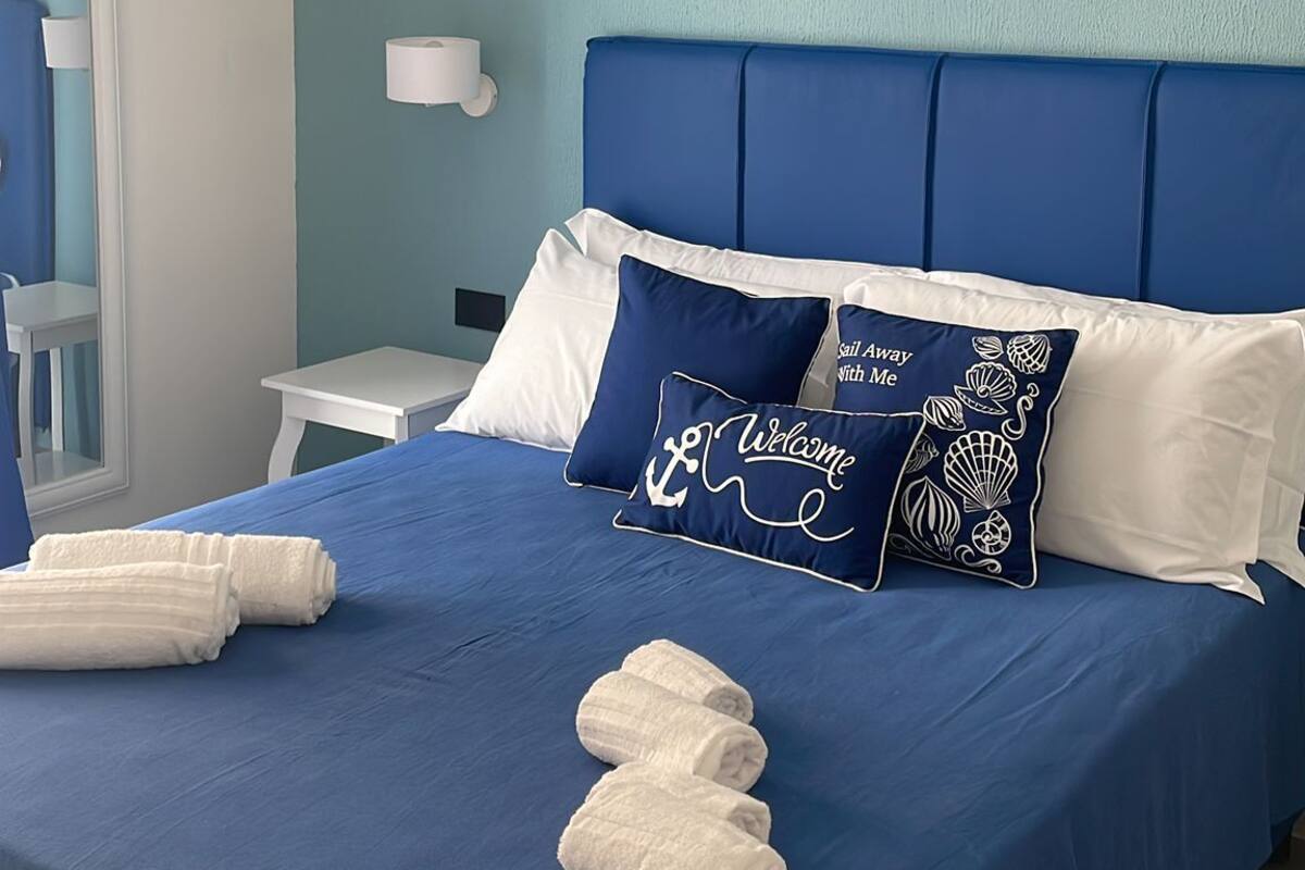 A neatly made bed with blue bedding, white pillows, and a decorative pillow with text. There is a white air conditioning unit on the wall, a small white table, and a white chair.