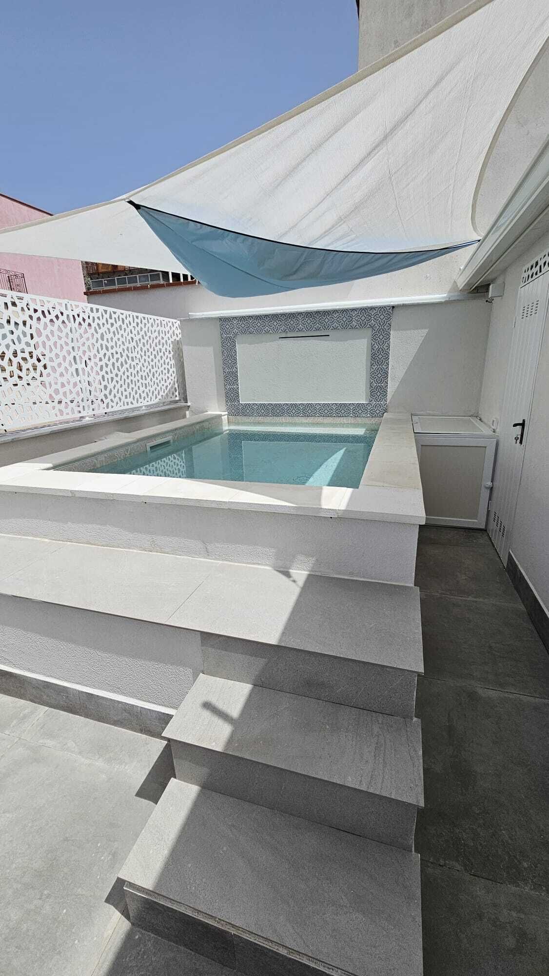 Ein moderner Außenpool mit integriertem Sitzbereich, einem überdachten Bereich mit blauem Sonnensegel und einer Wand mit Musterung.