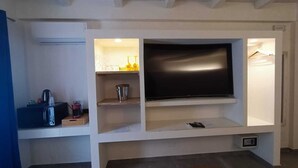 A modern living room with a flat-screen TV mounted on a white entertainment unit. There is a coffee maker and a kettle on the lower shelf, and a smartphone on the shelf above.