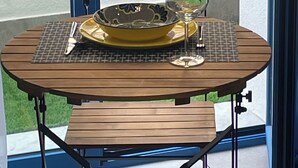 A round wooden table with a metal frame, a yellow plate, a wine glass, and a fork on a checkered placemat.