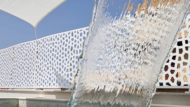 A water feature with a cascading waterfall in a pool area, surrounded by a white lattice fence and a clear blue sky.
