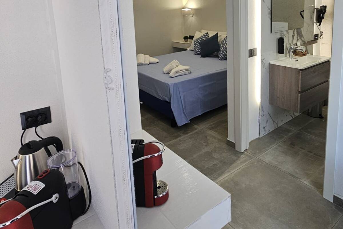 A modern hotel room with a large mirror, a bed with blue bedding, a bedside table with a kettle and a coffee machine, and a bathroom with a marble sink and mirror.