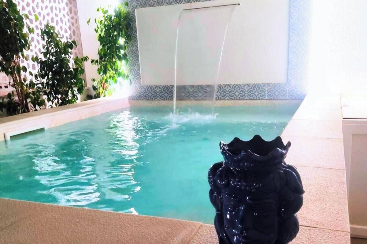 A pool with a waterfall feature, surrounded by plants and a tiled wall.