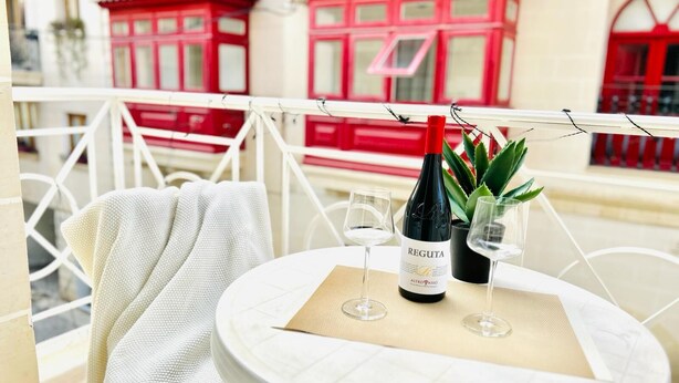 A round table with a bottle of wine, two glasses, and a potted plant on a balcony with white railings and red buildings in the background. - FUN GAME St. Julians Apartment 2 (St. Julians)