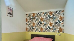 Ein Schlafzimmer mit einem Bett, einem Nachttisch, einer Wand mit tropischem Blattmuster, einem Oberlicht und einem gerahmten Bild.