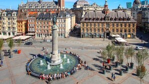 Ein öffentlicher Platz mit einem zentralen Brunnen, umgeben von historischen Gebäuden und Bäumen.