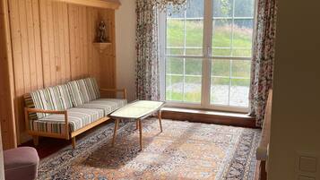 Une pièce avec des boiseries, un canapé, un tapis à motifs, une chaise en bois et une grande fenêtre avec des rideaux.