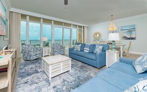 A spacious living room with a large window overlooking the sea, a blue sofa set, a coffee table, and a chandelier. - Phoenix Gulf Towers 706 2 Bedroom Condo by RedAwning (Orange Beach)