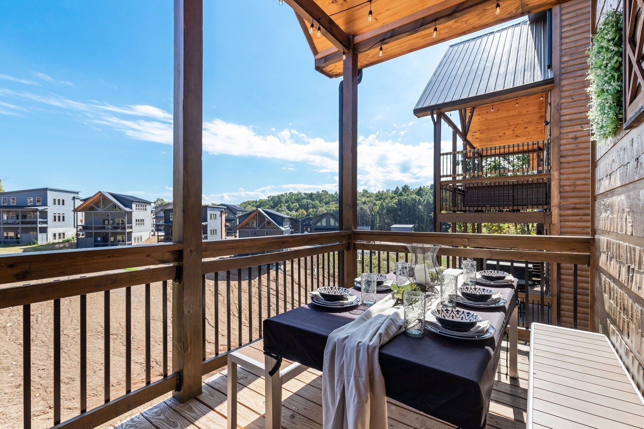 A wooden deck with a table set for dining, overlooking a residential area with houses and a forested hill.