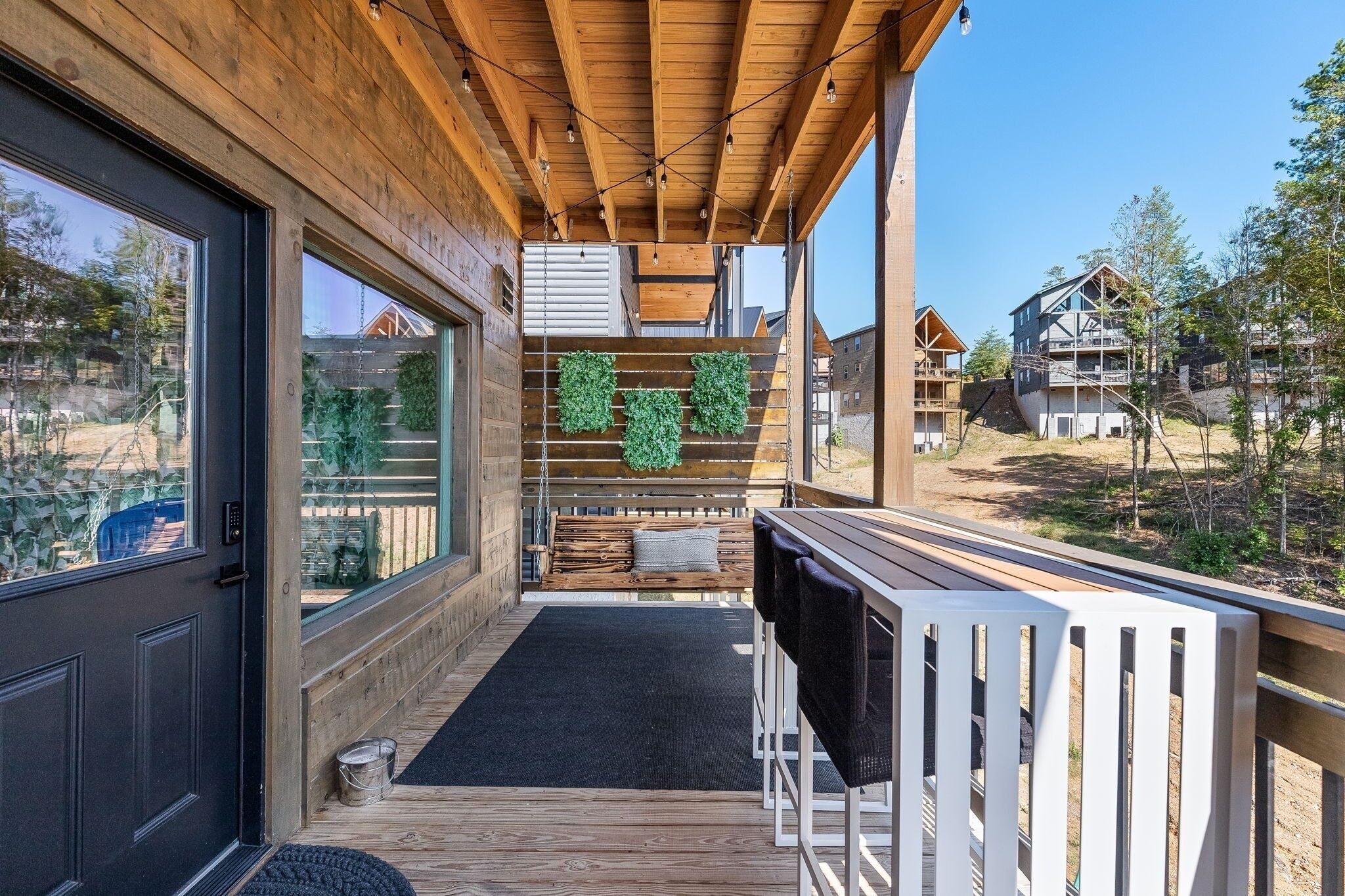 A wooden deck with a bench and a table, a door with a black mat, and a view of surrounding buildings.