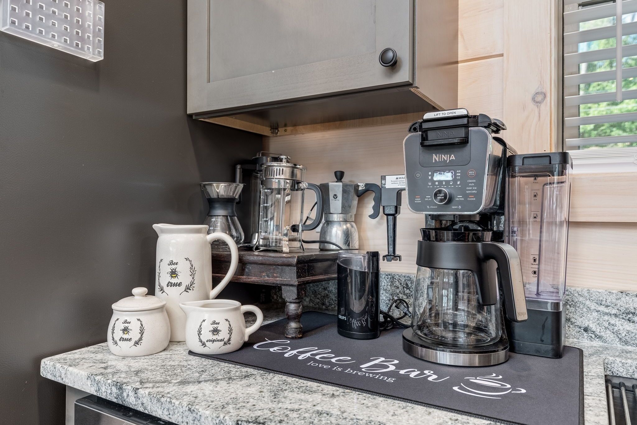 Un coin café équipé d’une machine à expresso Ninja, d’une cafetière à piston et de différentes tasses à café, le tout posé sur un comptoir en granit.