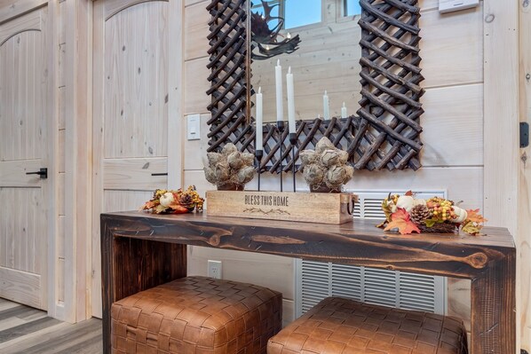 Un intérieur en bois, avec une console en bois foncé, deux poufs en cuir, un miroir orné d’un motif en treillis et une plaque portant l’inscription « QUE CETTE MAISON SOIT BÉNITE ».