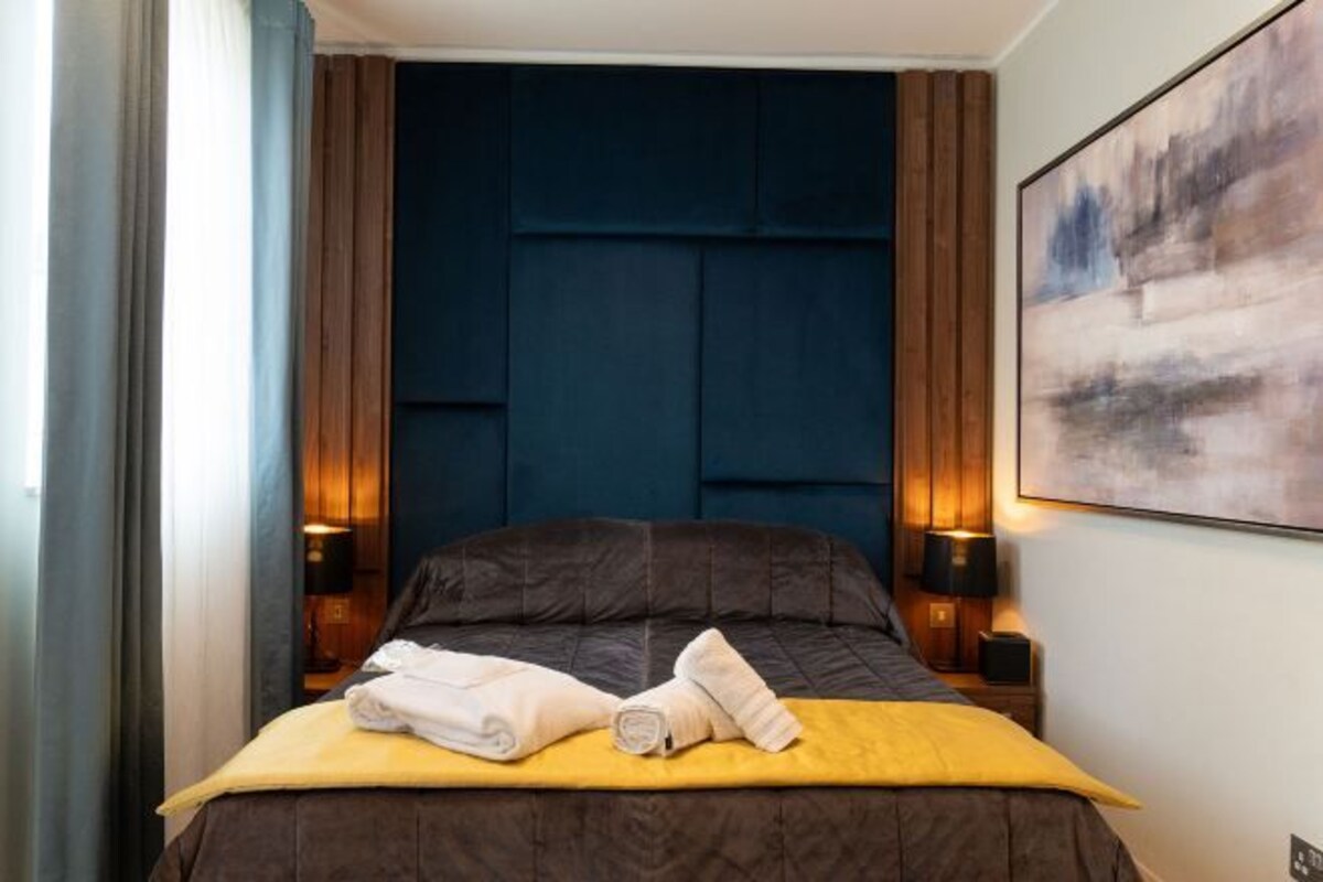 A hotel room with a dark blue headboard, a bed with a yellow bedspread, two white towels, and two bedside lamps.