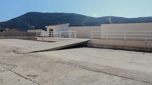 Una terraza en la azotea con suelo de hormigón, barandillas de metal y un pequeño edificio con techo rojo.