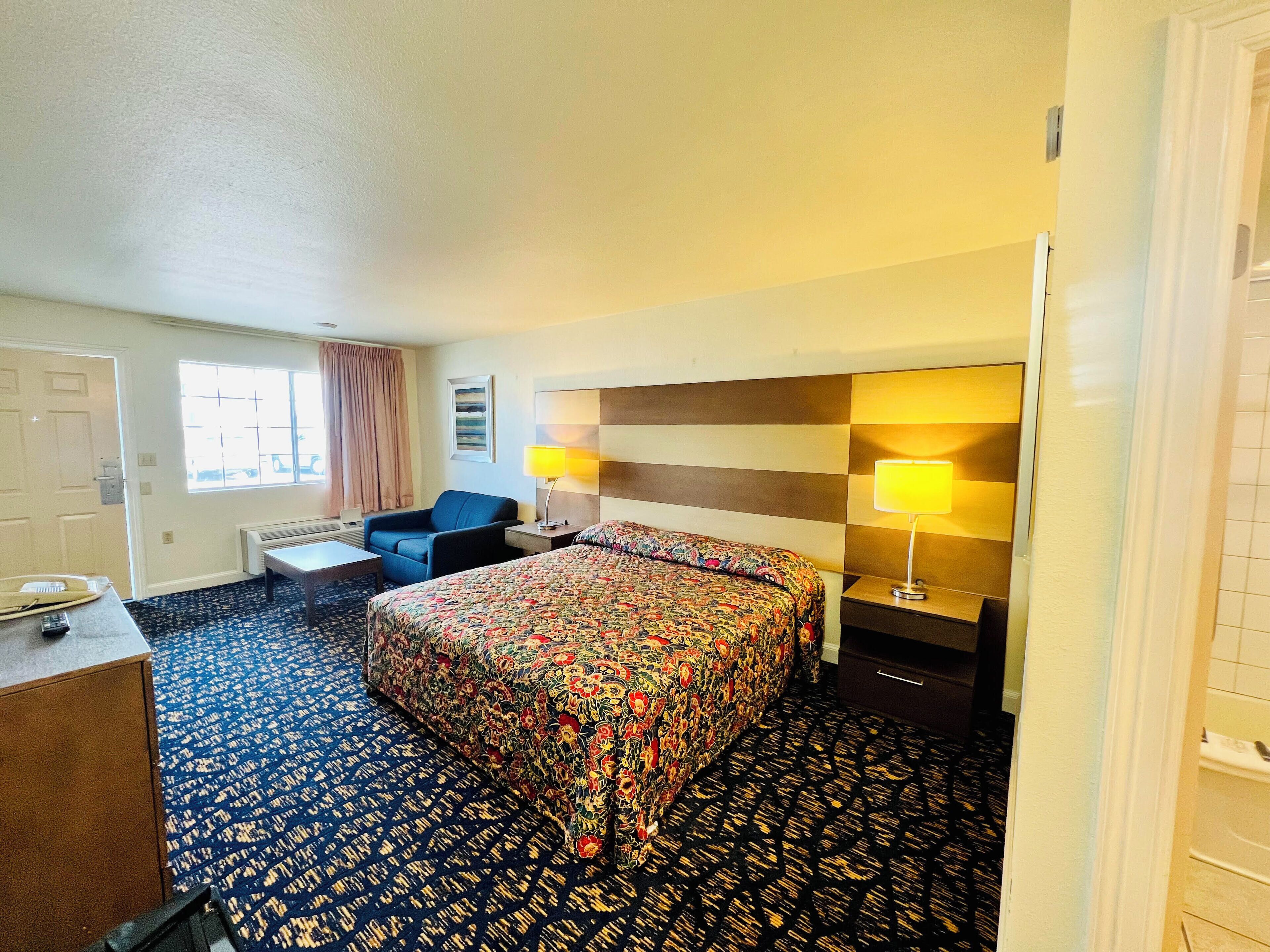 Habitación de hotel con cama, escritorio, silla, ventana con cortinas y cabecera con lámparas.