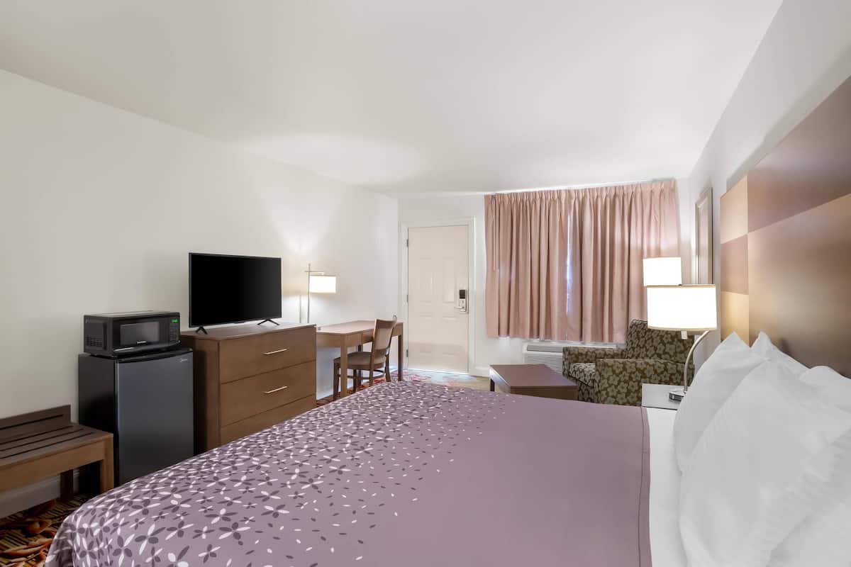 Hotellihuone, jossa on sänky, työpöytä, televisio, jääkaappi, tuoli ja ikkuna, jossa on verhot.