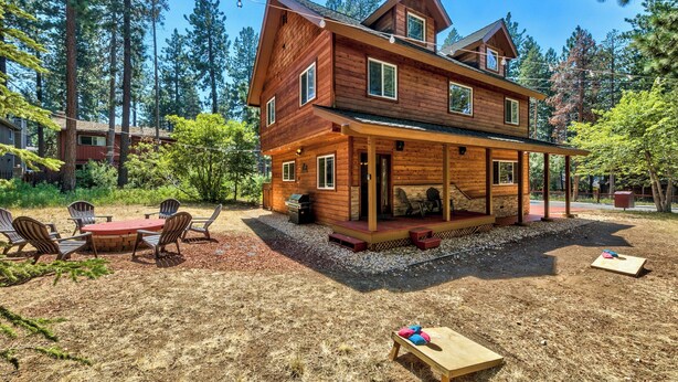 A two-story log cabin with a covered patio, outdoor seating, and a view of the surrounding forest.