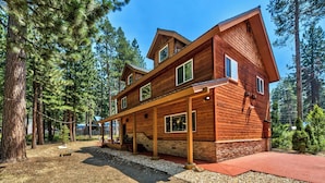 A two-story wooden cabin with a covered porch, surrounded by tall pine trees.