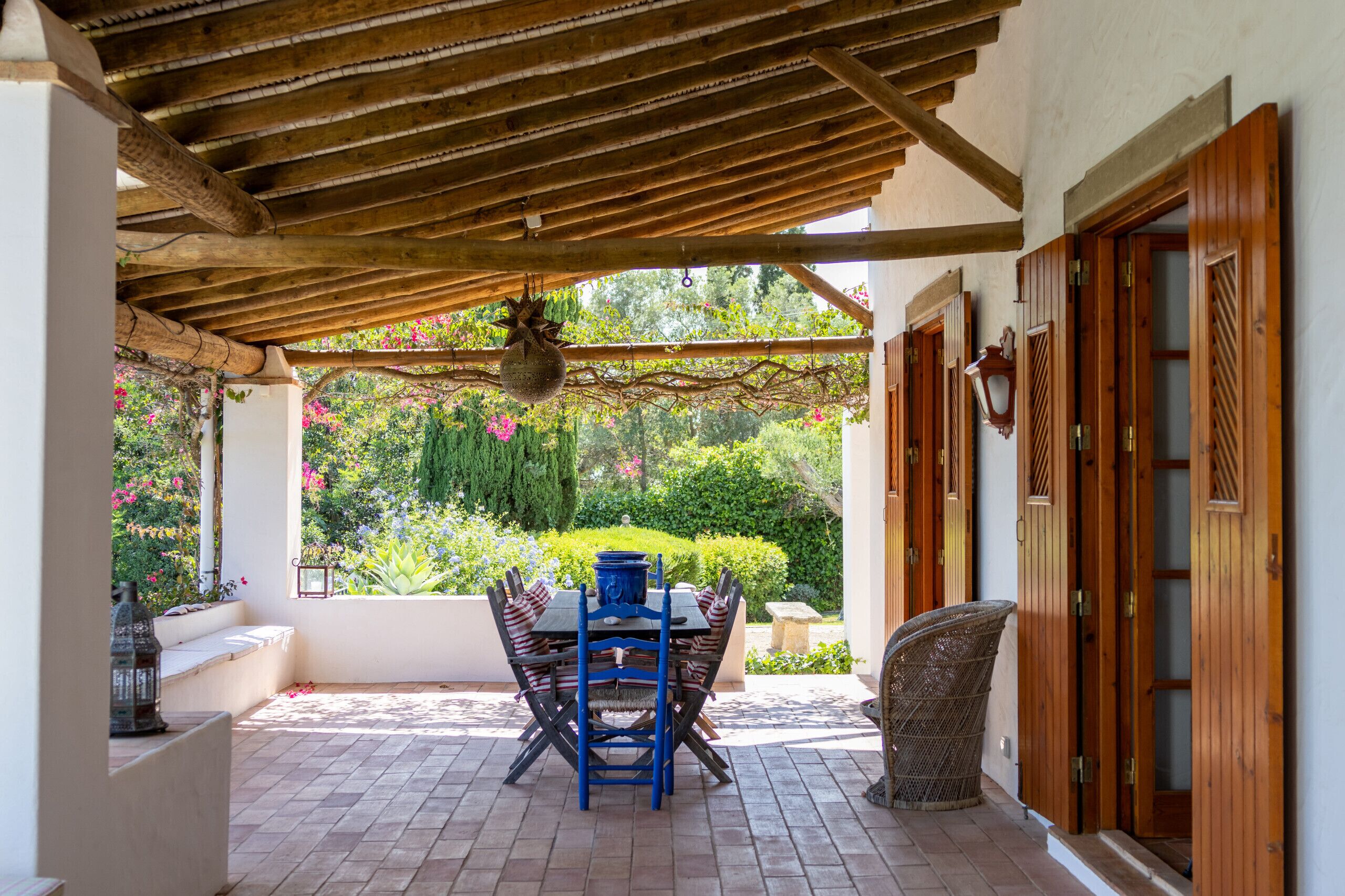 Une terrasse couverte avec une table et des chaises, une lanterne suspendue et une vue sur un jardin fleuri et verdoyant.