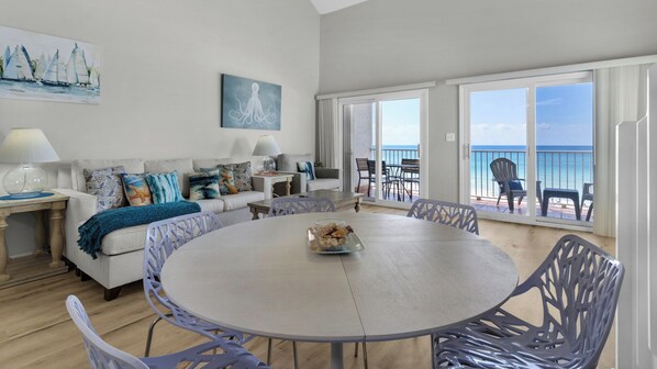 A spacious living area with a round table and chairs, a sofa with decorative pillows, a painting of sailboats, and a view of the ocean through large windows.