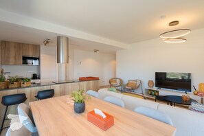 Eine moderne Küche mit einer zentralen Kochinsel, ein Essbereich mit Holztisch sowie ein Wohnzimmer mit Sofa und Fernseher.