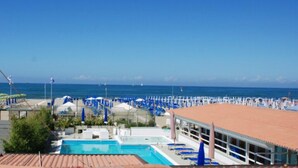 Een zwembad met ligbedden en parasols, een strand met ligbedden en parasols, en een heldere blauwe lucht.