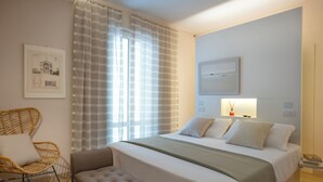 Een hotelkamer met een bed, een stoel, een bankje, een raam met gordijnen, een kunstwerk aan de muur en een nachtkastje met een lamp.