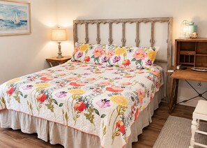 A bed with a floral bedspread, a wooden headboard, a bedside lamp, and a small wooden nightstand with a lamp and a decorative item. - 109 Georges Bay Road(Bermuda Bay) (Surfside Beach)
