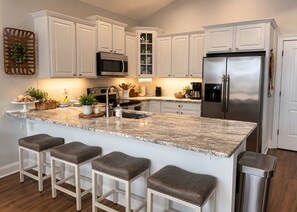 A modern kitchen with white cabinetry, a stainless steel refrigerator, a granite countertop, and bar stools. - 109 Georges Bay Road(Bermuda Bay) (Surfside Beach)
