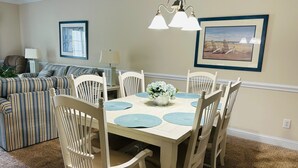 A dining area with a table set for six, surrounded by six white chairs with cushioned seats. There is a hanging light fixture above the table. - Villa #1608 2 bedroom 2 bath (Garden City)