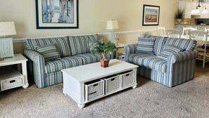 A living room with striped sofas, a white coffee table, and a dining area with white chairs. - Villa #1608 2 bedroom 2 bath (Garden City)