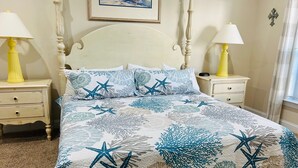 A bedroom with a bed featuring a starfish and coral pattern, two bedside tables with lamps, a framed picture on the wall, and a window with curtains. - Villa #1608 2 bedroom 2 bath (Garden City)