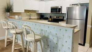 A kitchen with white cabinets, a stainless steel refrigerator, a microwave, and a stove. There are three wicker bar stools and a potted plant. - Villa #1608 2 bedroom 2 bath (Garden City)