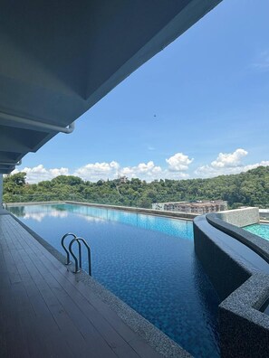 An infinity pool with a wooden deck, surrounded by lush greenery and a clear blue sky.