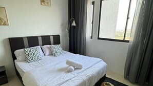 Ein ordentlich bezogenes Bett mit weißer Decke und Kissen, ein schwarzes Kopfteil, ein Fenster mit Vorhängen und ein Teppich mit Blumenmuster.