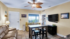 A compact living space with a sofa, a dining table with stools, a ceiling fan, a TV, and a window with blinds. - Pool, Marinas & Waterfront Dining Nearby @ The Waterway 403 (Wrightsville Beach)