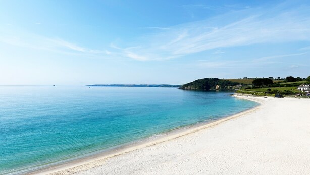 Une plage avec une eau d’un bleu limpide, un rivage sablonneux et une petite île verdoyante, avec des bâtiments visibles au loin.