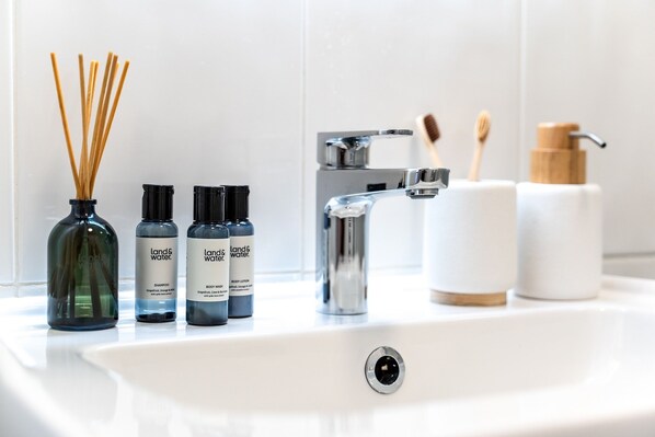 A bathroom sink with three bottles of shampoo, a body wash, and a body lotion. There are also two toothbrushes and a wooden-handled toothbrush holder.