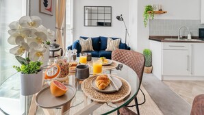 Une cuisine moderne avec des meubles blancs, un évier et un plan de travail. On y trouve un coin repas avec une table en verre, des assiettes et un demi-pamplemousse.