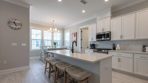 Una cocina moderna con armarios blancos, una isla central con taburetes y un microondas de acero inoxidable, además de un reloj en la pared.