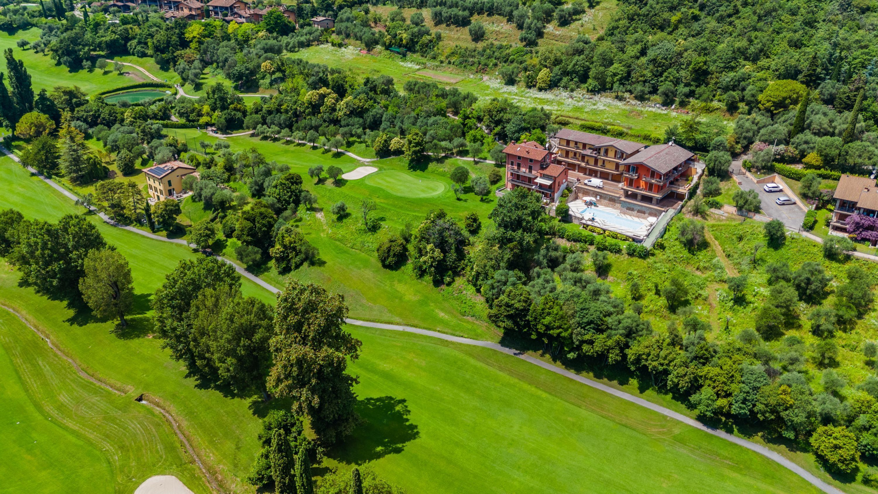 An aerial view of a large, multi-story building with a swimming pool, surrounded by lush green fields and trees.